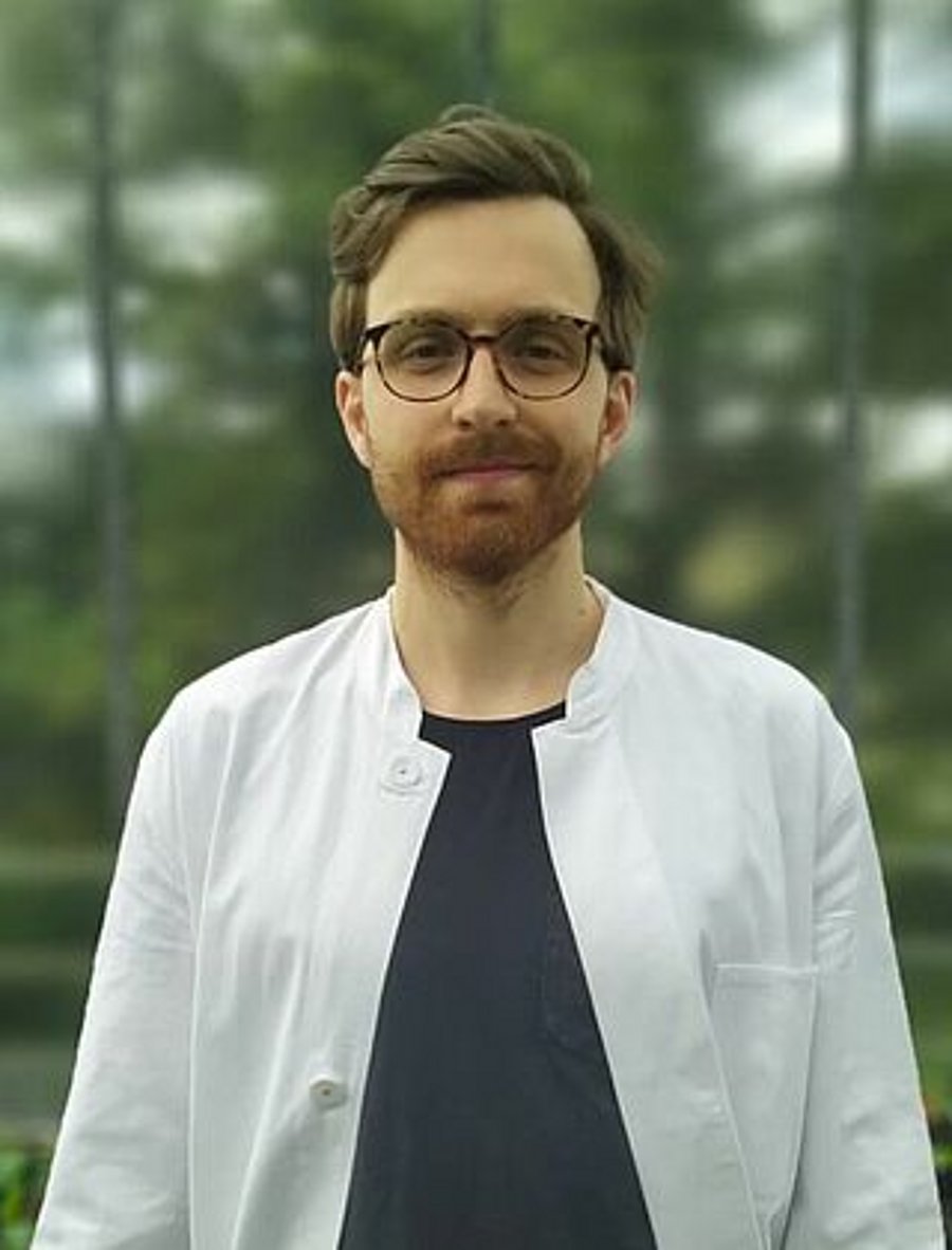 A photo of Lukas Beule. It shows a man with brown hair and a beard looking directly into the camera. He smiles slightly. He wear a white shirt over a black t-shirt. He stands in front of trees. 