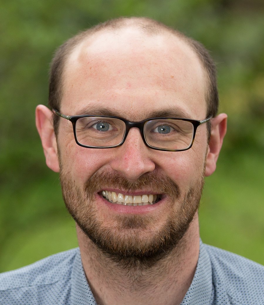 A photo of Jan Peters. It shows a balding, smiling man in his thirties. He has brown hair, a beard, and blue eyes. He wears glasses and a light blue shirt. The background of the photo is green and blurred.
