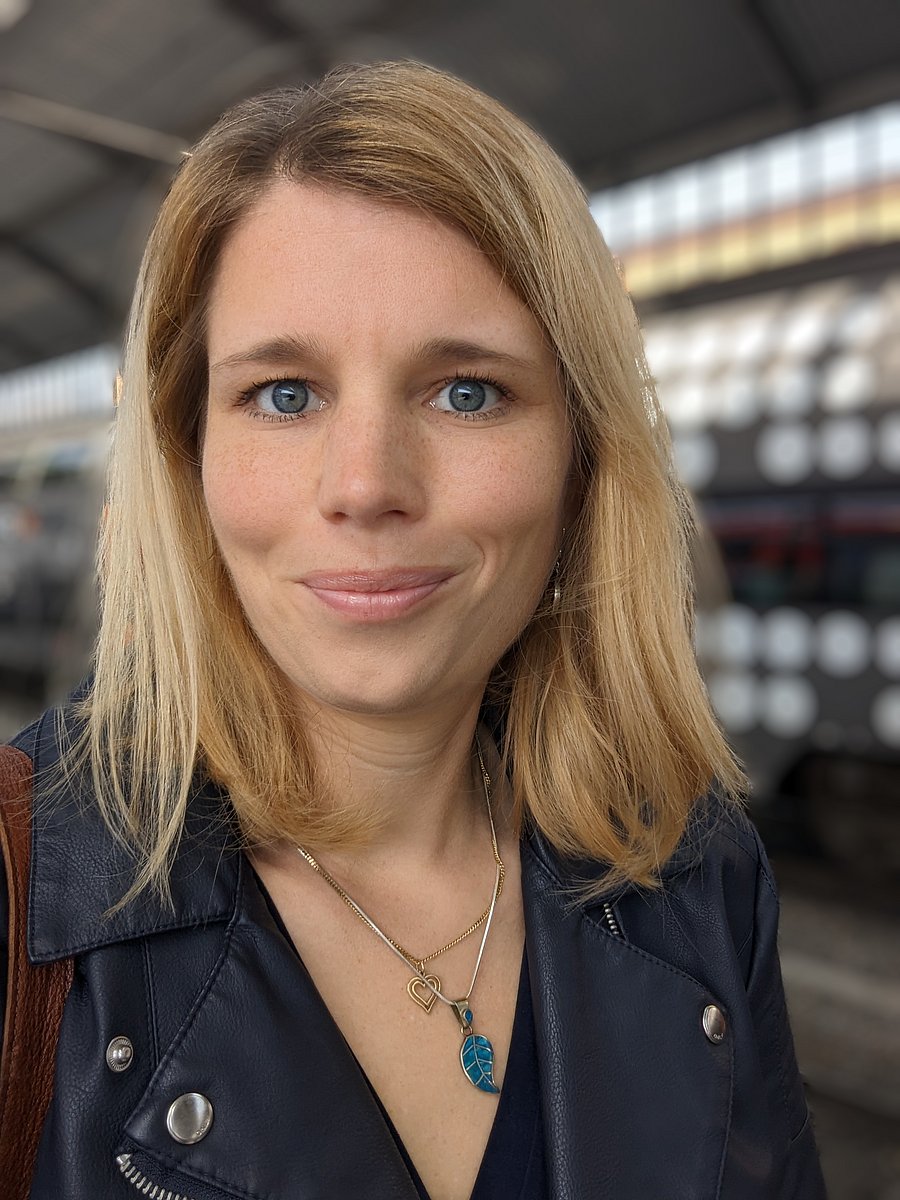 A photo of Linda Homann. It depicts a smiling woman in her late thirties, with blue eyes and blonde hair. She is wearing a dark blue leather jacket over a black shirt. The strap of a brown leather bag is visible over her right shoulder. A train station hall can be seen in the background.